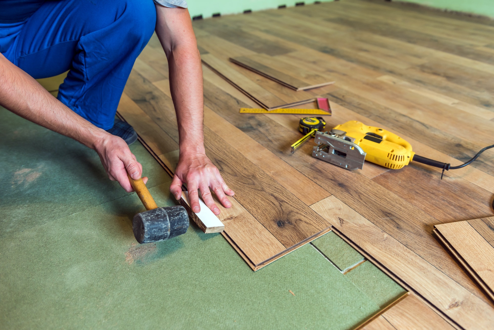 Installer fitting laminate flooring with a mallet and tools over underlayment.